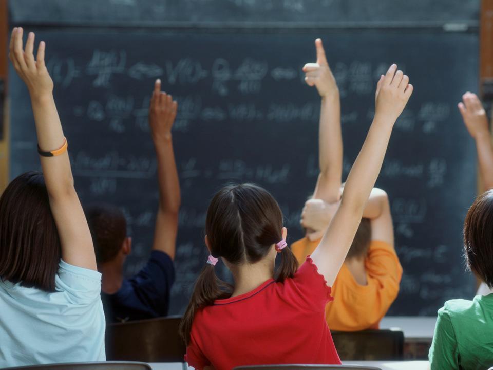 children facing a blackboard in a classroom, all withtheir hands raised.