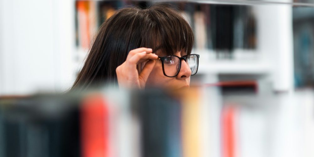 girl with glasses  peering over the top of books on a book shelf