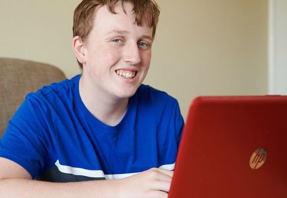 A smiling teenage boy sat at a desk with a open laptop