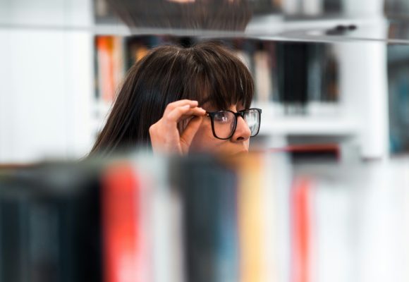 girl with glasses  peering over the top of books on a book shelf