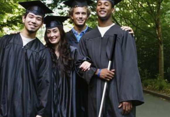 Four smiling students in graduation gowns and caps. The student on the far right is holding a long cane.