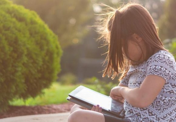 young girl sat outside in the sunshine looking down at a tablet she is holding